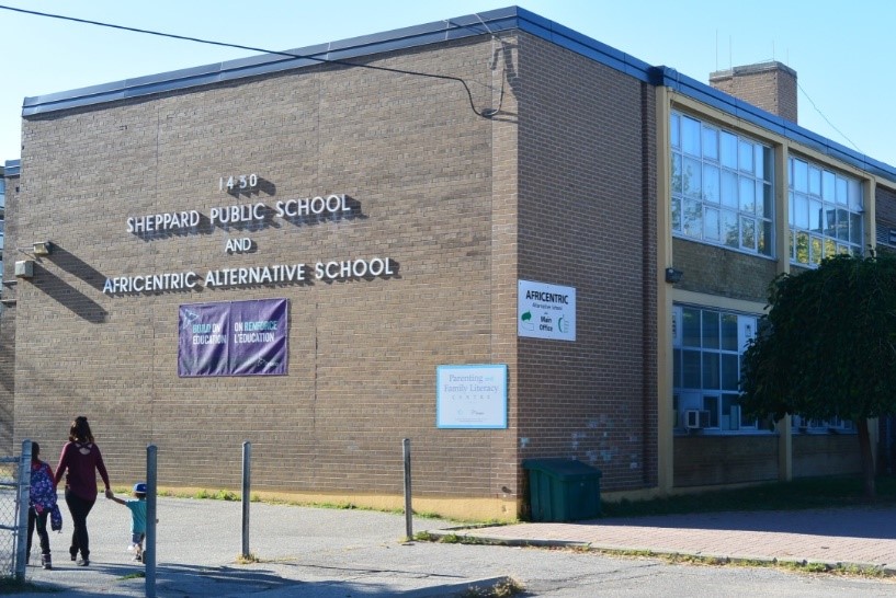 Backsteinbau der Sheppard Public School mit Schriftzug ‚Africentric Alternative School‘; im Vordergrund gehen eine Frau und zwei Kinder durch das Schultor.