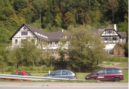 Fachwerkgebäude eines Gefängnisschul-Standorts in bewaldeter Hanglage; vor dem umzäunten Areal verläuft eine Landstraße mit geparkten Autos.